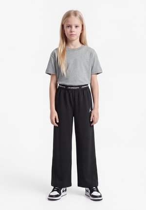 Jeune fille aux longs cheveux blonds porte un t-shirt gris, un pantalon Jordan noir et des baskets noires et blanches, debout devant un fond blanc.