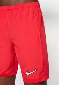 Røde Nike-shorts laget av lett materiale, med elastisk midjebånd, sidelommer og en hvit brodert logo nederst til venstre.