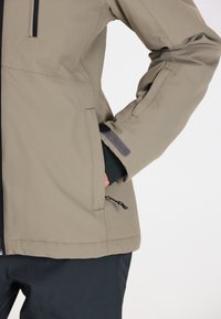 Ljusbrun, vattentät jacka med en slät textur, dragkedjeficka och justerbara ärmmuddar med kardborre och dragsnöre.