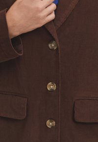 Blazer de pana marrón con dos bolsillos en el pecho, tres botones redondos y un acabado texturizado. Mano agarrando la solapa.