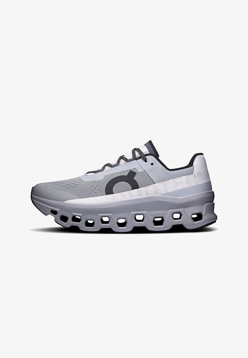 Scarpa sportiva grigia con tomaia in mesh, design della suola intermedia esagonale e accenti neri. Presenta lacci testurizzati e un logo prominente sul lato.