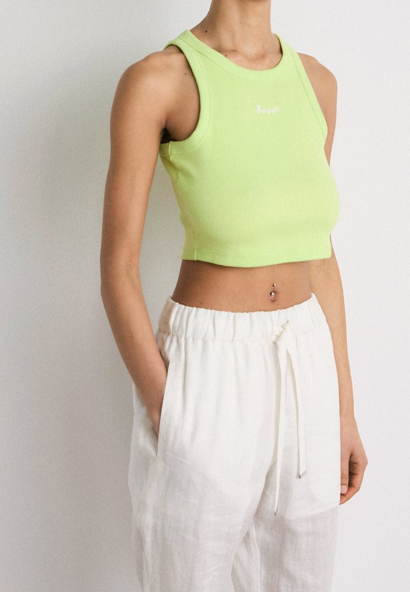 Femme portant un crop top sans manches vert clair et un pantalon blanc ample, mains dans les poches, montrant un piercing au nombril.