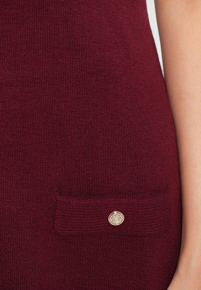Burgundy knit fabric with a ribbed texture, featuring a small patch pocket and a circular gold accent button.