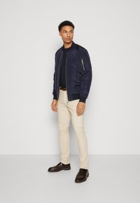 Chaqueta bomber de marina con un bolsillo con cremallera, pantalones beiges y zapatos marrón oscuro. Diseño sencillo, textura suave y corte ajustado.
