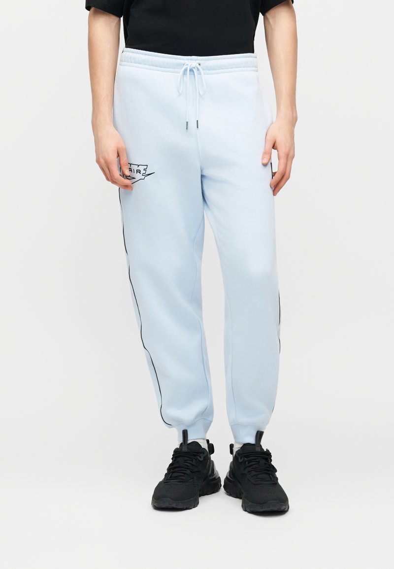 Persona che indossa pantaloni sportivi Nike Air azzurri chiaro con strisce nere ai lati e scarpe da ginnastica nere, su sfondo bianco uniforme.