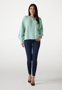 Mintgrüner Strickcardigan mit einem strukturierten Muster, Rundhalsausschnitt und fünf Knöpfen an der Vorderseite. Kombiniert mit dunkelblauen Skinny-Jeans.