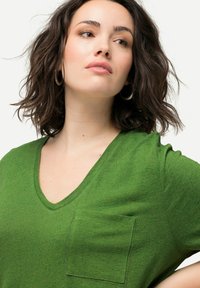 Camiseta verde de cuello en V hecha de tela suave, con un bolsillo parche y detalles de costura sutiles. Manga corta y corte holgado.