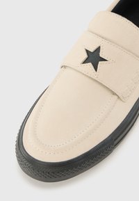 Beige nubuck slip-on sko med en svart stjerneskjæring, gummisåle med teksturert mønster, og sydd detalj rundt kanten.