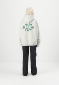 Light gray hoodie with "Replay Breakfast Crew" printed in dark green on the back. Paired with dark denim pants. Soft fabric, loose fit.