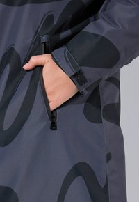 Giacca resistente all'acqua realizzata in tessuto grigio scuro con motivi astratti neri, tasche laterali con zip e dettagli ai polsini in Velcro.
