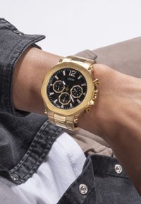 Auksinės spalvos "Guess" laikrodis su juodu ciferblatu ir trimis šešiakampiais subdials nešamas žmogaus ant riešo, kuris vilki tamsiai mėlyną džinsinį švarką ir baltą marškinį.