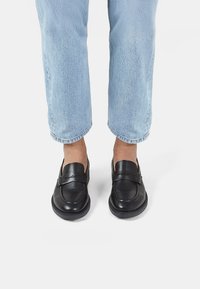 Mocasines de cuero negro con un acabado suave, que presentan una puntera cosida y una correa decorativa, combinados con jeans de tiro alto en azul claro con dobladillos.