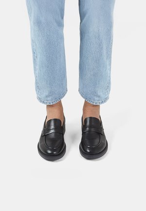 Mocasines de cuero negro con un acabado suave, que presentan una puntera cosida y una correa decorativa, combinados con jeans de tiro alto en azul claro con dobladillos.