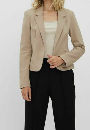 Vrouw draagt een beige op maat gemaakte blazer over een crème ribgebreide tanktop en zwarte wijde broek met hoge taille, staand tegen een effen achtergrond.