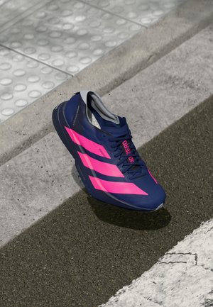 Chaussure de course bleu marine avec des rayures rose vif et texte "40 ZERO" sur l'asphalte près du bord du trottoir et du pavage tactile.