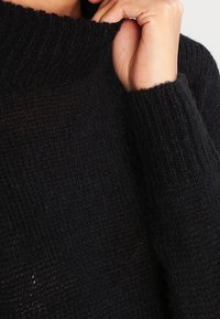 Musta neulepusero, jossa on korkea ribattu kaulus ja teksturoitu kangas. Ominaisuuksina pitkät hihat, joissa on selkeästi määritellyt hihansuut.