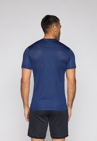 Marineblå træningsskjorte med korte ærmer, lavet af tekstureret stof, har rund hals og diskrete skulderdetaljer. Bæret med sorte shorts.