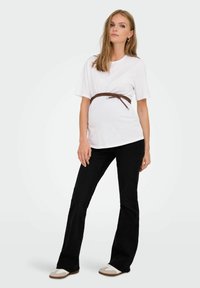 White cotton t-shirt with short sleeves, belted in brown leather, paired with black flared jeans and beige sneakers, standing on a neutral background.