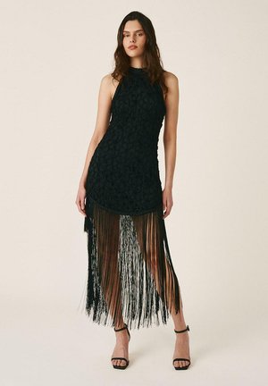 Mujer con vestido negro sin mangas, cuerpo con textura y falda larga con flecos, combinada con sandalias negras de tacón alto, de pie.