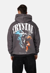 Felpa grigia con cappuccio, caratterizzata da una grande grafica di un leone in blu e arancione, con la dicitura "CRYSTAL PARIS" stampata sopra. Realizzata in un tessuto morbido con una vestibilità comoda.