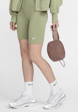 Calções verdes em mistura de algodão com um logótipo branco da Nike, combinados com ténis brancos da Nike e meias caneladas. Bolsa castanha com fecho, segurada na mão.