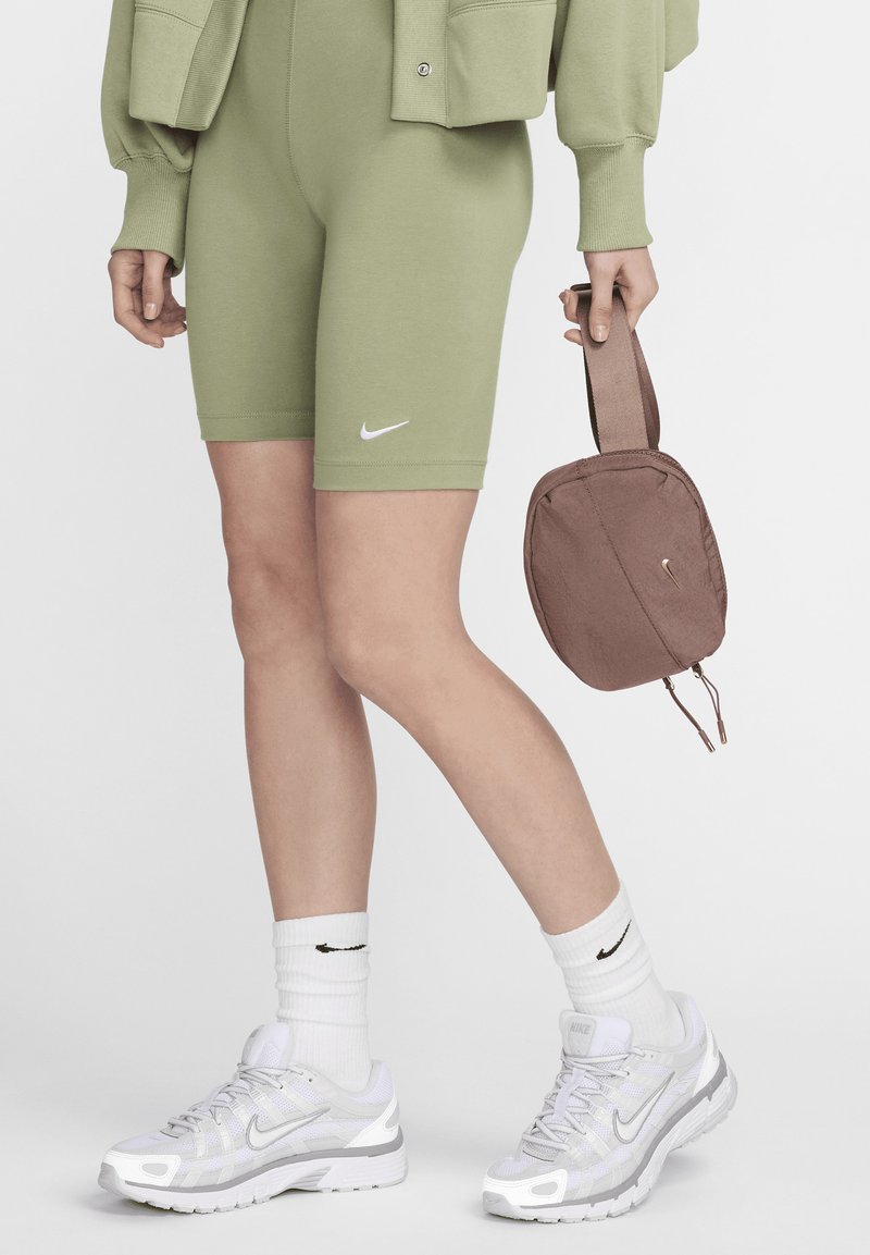 Zöld pamut keverék sort fehér Nike logóval, fehér Nike cipőkkel és bordás zoknikkal párosítva. Barna, cipzáras tokot tart a kezében.