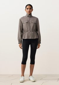Femme debout portant une veste grise zippée avec taille à cordon, des leggings noirs courts et des baskets blanches sur un sol carrelé pâle.