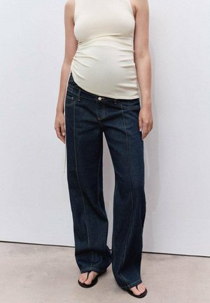 Pregnant person wearing a white sleeveless top, dark wide-leg jeans, and black flip-flops standing against a plain wall.