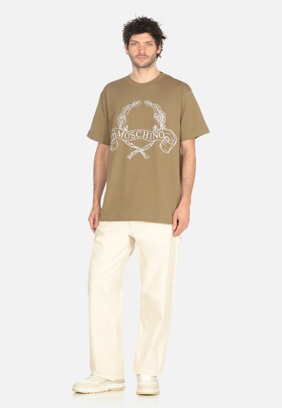 Uomo che indossa una t-shirt marrone Moschino, pantaloni a gamba larga color crema e sneakers beige, in piedi davanti a uno sfondo bianco semplice.