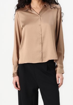 Vrouw draagt een beige satijnen blouse met knopen en lange mouwen en een zwarte broek met hoge taille tegen een witte achtergrond.