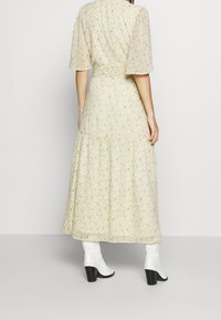 Svetle žluté maxi šaty s květinovým vzorem, krátkými puffovými rukávy, stupňovitou spodní částí a stylizované s bílými kojicími botami.