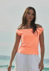 Top naranja neón sin hombros con mangas cortas, hecho de una tela ligera, combinado con pantalones cortos blancos de corte holgado.