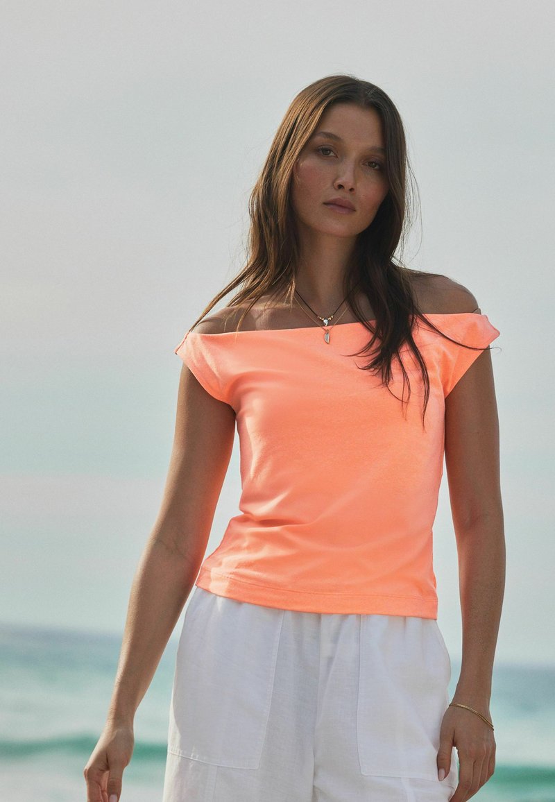 Top naranja neón sin hombros con mangas cortas, hecho de una tela ligera, combinado con pantalones cortos blancos de corte holgado.