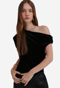 Černý sametový top s odhalenými rameny a krátkými rukávy, s jemnou texturou a splývavým střihem. Modelka má na sobě tmavé kalhoty s vzorem.