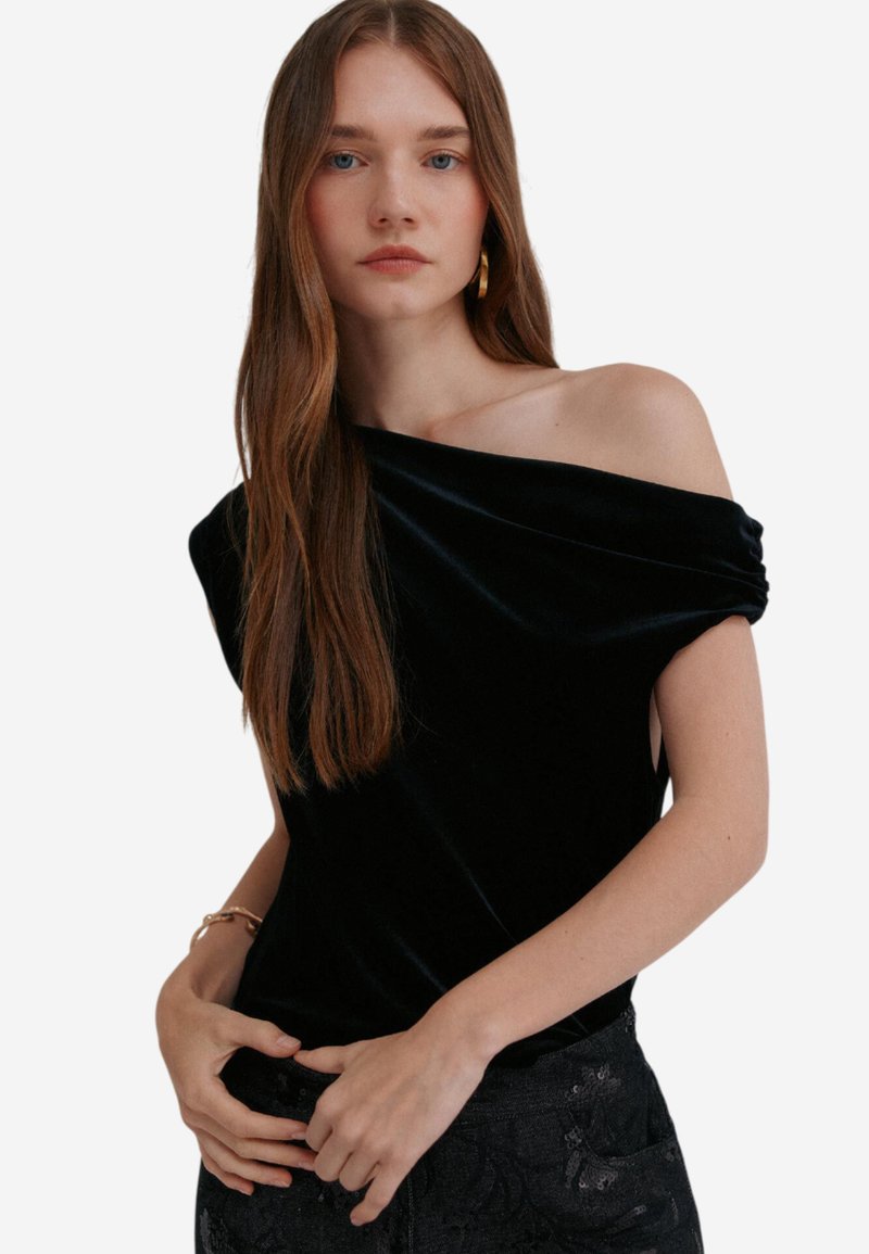 Černý sametový top s odhalenými rameny a krátkými rukávy, s jemnou texturou a splývavým střihem. Modelka má na sobě tmavé kalhoty s vzorem.