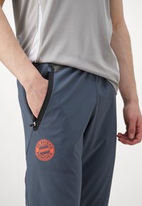 Grå sportbukser med et orange FC Bayern München-logo, lynlåslommer på siden og en let, glat stoftekstur.