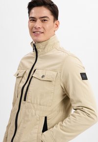 Beige Zip-Jacke mit Stehkragen, zwei Brusttaschen, seitlichen Taschen und Schulterpatch. Glattes Gewebe mit taillierter Passform.