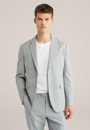 Fiatal férfi világosszürke öltönyben és fehér pólóban, egyik kezét zsebbe dugva áll, sima bézs háttér előtt.