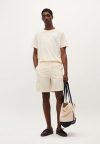 Tricou din bumbac de culoare deschisă cu imprimeu albastru, asociat cu pantaloni scurți cargo bej. Modelul ține o geantă tip tote în două nuanțe, cu accente albastre.