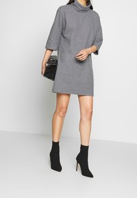 Vestito grigio a collo alto con maniche corte, vestibilità morbida e texture liscia; abbinato a stivaletti neri e una borsa nera.