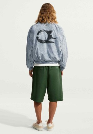 Person med skulderlangt hår iført en grå jakke med sort grafik og mørkegrønne shorts, stående med ryggen til mod en lys baggrund.