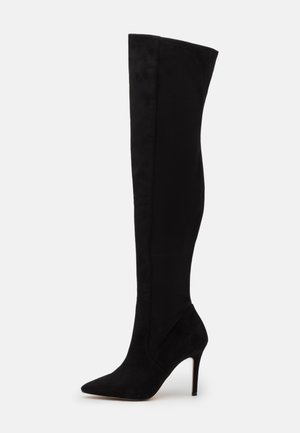 Bota de tacón stiletto negra por encima de la rodilla, con punta afilada y material de ante suave, exhibida sobre un fondo blanco.