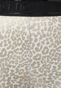 Goldene Leggings mit Leopardenmuster auf weißem Grund, ausgestattet mit einer glatten Textur und einem schwarzen elastischen Bund mit geprägtem Branding.