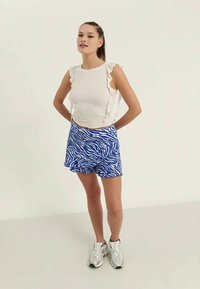 Top blanco con mangas con volantes combinado con shorts de estampado de cebra en azul y blanco. La modelo lleva zapatillas blancas y está de pie sobre un fondo claro.