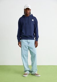 Navy blue hoodie with kangaroo pocket, paired with light blue baggy jeans and brown sneakers. White cap with "DICKIES" logo. Green floor backdrop.