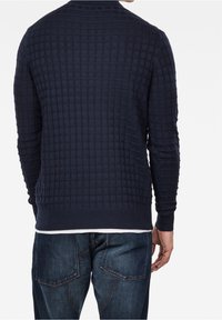 Mörkblå stickad tröja med en strukturerad rutmönster. Rund halsringning och ribbade ärmslut och nederkant, tillsammans med blå denimjeans.