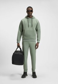 Groene sportieve hoodie met een voorzak, gecombineerd met bijpassende slim-fit broek. Zwarte sporttas, donkere sneakers en een zonnebril zijn te zien.