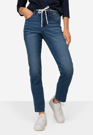 Jeans in denim di un blu medio, con una vestibilità classica, design a cinque tasche e un laccio bianco in vita. Indossati con sneakers bianche.