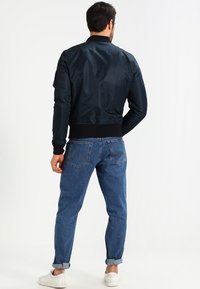 Homme portant une veste bomber noire, un jean bleu retroussé et des baskets blanches, se tenant de dos contre un fond clair.