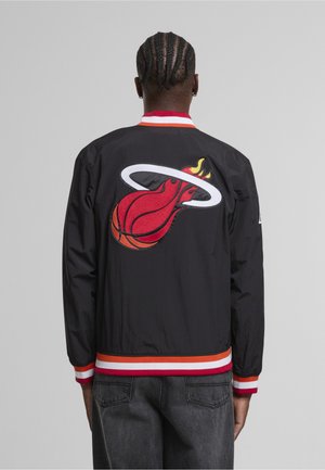 Homme aux cheveux tressés portant une veste noire Miami Heat avec un logo de ballon de basket en flammes dans le dos, associé à un jean sombre, vu de dos.